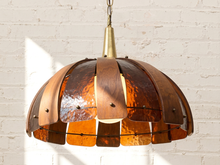 Load image into Gallery viewer, A mid-century modern pendant light with a dome shade made of overlapping hammered copper and amber glass panels, suspended against a white brick wall.

