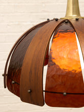 Load image into Gallery viewer, A close-up of a hanging pendant light with curved wooden slats and a glowing orange glass shade, suspended from a brass-toned ceiling mount against a white brick wall.
