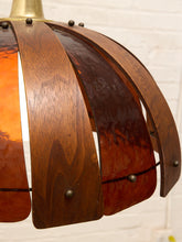 Load image into Gallery viewer, A close-up of a vintage-style pendant lamp shade featuring curved wooden slats with a warm amber-orange glass or metal backing, secured by small metal rivets, against a white brick wall background.
