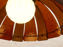 Load image into Gallery viewer, A close-up of a modern pendant light with a glowing white globe bulb and a curved shade made of glossy, warm brown wood slats, set against a white brick wall.
