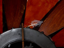 Load image into Gallery viewer, Close-up of a vintage wooden ship's wheel with dark brown wood spokes, a black metal rim, and a rusted brown center hub against a red background.
