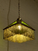 Load image into Gallery viewer, A vintage-style hanging pendant light with a geometric frame, green glass panels, and a long fringe of gold-toned tinsel-like strands, glowing warmly against a white brick wall.
