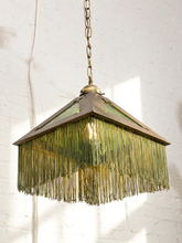Load image into Gallery viewer, A vintage-style hanging pendant light with a bronze-toned metal frame, green-tinted glass panels, and a long fringe of hanging metallic strands, shown against a white brick wall.
