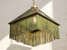 Load image into Gallery viewer, A vintage-style square pendant light with a bronze metal frame, green-tinted glass panels, and long hanging fringe, shown hanging against a white brick wall.
