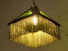 Load image into Gallery viewer, A hanging pendant light with a dark metal frame, green glass panels near the top, and a long fringe of gold-toned tinsel or metallic strands below, glowing warm yellow from an exposed bulb.
