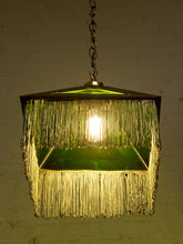 Load image into Gallery viewer, A vintage-style hanging pendant lamp with a green glass shade, gold-toned metal frame, and long fringe trim, illuminated against a warm off-white wall.
