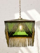 Load image into Gallery viewer, A hanging pendant light with a geometric dark metal frame, green glass panels, and long fringe in dark green and gold-toned threads, suspended against a white brick wall.
