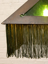 Load image into Gallery viewer, A close-up of a decorative hanging lamp or light fixture with a triangular dark metal frame, green glass panels, and long dark fringe tassels against a light brick wall.
