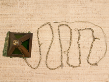 Load image into Gallery viewer, A vintage brass chain with a square wooden stopper featuring green glass panels and a brass grommet, laid out on a beige woven fabric background.
