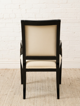 Load image into Gallery viewer, A high-back dining chair with a glossy black frame and cream-colored upholstered seat and backrest, positioned against a white brick wall and light beige rug.
