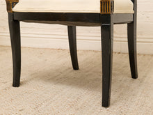 Load image into Gallery viewer, Close-up of a black wooden stool or bench with a cream upholstered seat and gold-toned decorative corner accents, shown on a woven beige rug against a white wall.
