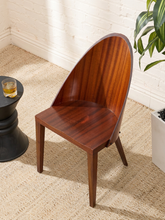 Load image into Gallery viewer, A mid-century modern wooden dining chair with a curved backrest and tapered legs, shown on a woven jute rug in a bright room with a white brick wall, a potted plant, and a small side table.
