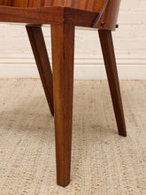 Load image into Gallery viewer, Close-up of a wooden chair leg with a rich medium-brown finish, resting on a textured beige woven rug against a white brick wall.
