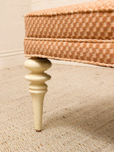 Load image into Gallery viewer, Close-up of an upholstered bench or ottoman with a woven peach and beige patterned fabric and turned cream-colored legs, shown on a textured beige rug.
