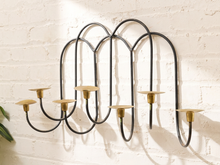 Load image into Gallery viewer, A black metal wall-mounted candelabra with five candle holders and brass cup holders, mounted on a white brick wall.
