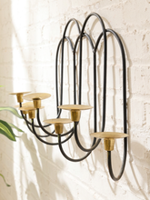 Load image into Gallery viewer, A black metal multi-arm wall sconce with brass candle cups, mounted on a white brick wall with soft sunlight and shadows.

