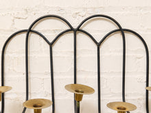 Load image into Gallery viewer, Black metal arched wall-hanging candle holder with brass candle cups, shown against a white painted brick wall.
