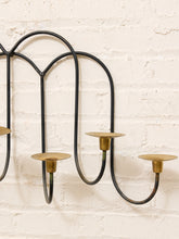 Load image into Gallery viewer, A close-up of a black metal wall-mounted candelabra with four candle cups in a brass finish, mounted on a white painted brick wall.
