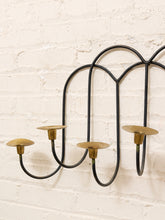 Load image into Gallery viewer, A black metal wall-mounted candelabra with five candle holders and brass-colored cups, mounted on a white painted brick wall.
