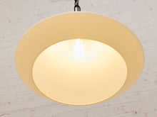 Load image into Gallery viewer, A modern circular pendant light with a wide beige outer shade and a warm cream inner diffuser, glowing with a bright bulb inside against a white brick wall background.
