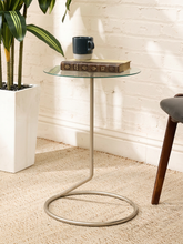 Load image into Gallery viewer, A modern round glass-top side table with a curved metal pedestal base, styled in a bright room with a potted plant, books, and a mug on top.
