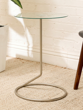 Load image into Gallery viewer, A modern round glass-top side table with a curved metal base, shown in a styled room setting on a textured beige rug against a white brick wall.
