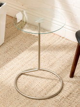 Load image into Gallery viewer, A modern round glass-top side table with a slim silver metal pedestal base and circular foot ring, shown on a woven beige rug in a bright room.
