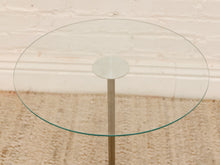 Load image into Gallery viewer, A round glass-top side table with a slim metallic stand, set against a light brick wall and textured beige rug.
