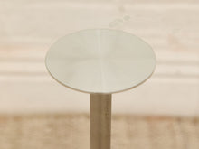 Load image into Gallery viewer, A minimalist round glass side table with a clear circular top and a slim beige metal pedestal base, set against a softly blurred beige background.
