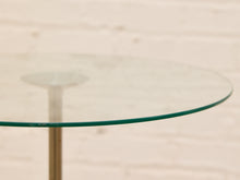 Load image into Gallery viewer, A close-up of a round clear glass tabletop with a thin green-tinted edge, supported by a slim metallic stand, set against a softly blurred light beige brick wall.
