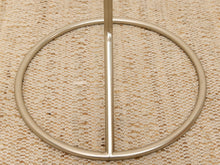 Load image into Gallery viewer, A close-up of a metallic gold circular ring stand or frame base resting on a textured beige woven rug.
