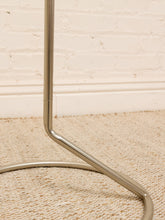 Load image into Gallery viewer, A close-up of a polished silver metal stand or frame leg resting on a beige woven rug against a white brick wall.
