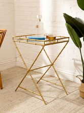 Load image into Gallery viewer, A gold metal side table with a bamboo-inspired frame design and a clear glass tray, styled in a bright room with a white brick wall, beige rug, and a green plant.
