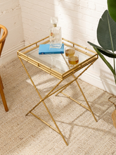 Load image into Gallery viewer, A gold metal side table with a clear glass top, styled with a blue book, a glass decanter, and two glasses in a bright room with a white brick wall and woven rug.
