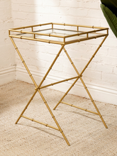 Load image into Gallery viewer, A gold-finished metal side table with a bamboo-inspired frame design, featuring a clear glass top and an X-shaped folding base, set on a beige rug against a white brick wall.
