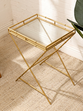 Load image into Gallery viewer, A square side table with a clear glass top and a gold metal frame designed to look like bamboo, shown on a woven beige rug against a white brick wall.
