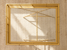 Load image into Gallery viewer, A gold-framed mirror with a bamboo-style border and a clear glass surface, shown against a textured beige woven background.

