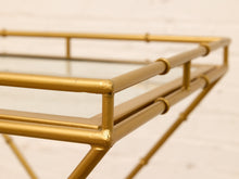 Load image into Gallery viewer, Close-up of a gold metal tray table with a glass top, showing the bamboo-inspired jointed frame against a light off-white brick wall background.
