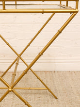 Load image into Gallery viewer, Close-up of a gold-finished metal side table with a bamboo-inspired frame design and a glass shelf, set against a white brick wall and light beige rug.
