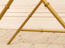 Load image into Gallery viewer, Close-up of a gold metal frame structure with a bamboo-inspired joint design, resting on a textured beige rug against a white paneled wall.
