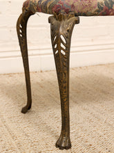Load image into Gallery viewer, Close-up of an antique-style chair leg with a carved bronze-toned metal foot and a floral patterned fabric seat cushion, set against a white brick wall and beige rug.
