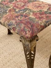 Load image into Gallery viewer, Close-up of an upholstered table or stool corner with a floral tapestry fabric in pink, red, blue, and beige tones, supported by an ornate carved bronze-toned leg on a textured cream rug.
