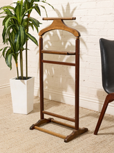 Load image into Gallery viewer, A wooden hanger rack with a curved top bar and three horizontal support bars, shown standing on a textured beige rug against a white brick wall. A potted green plant is on the left, and the corner of a black chair is on the right.
