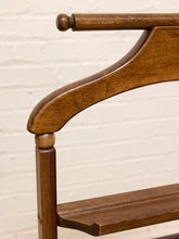 Load image into Gallery viewer, Close-up of a wooden coat rack with a warm medium-brown finish, featuring a curved top bar, turned round finials, and a lower shelf, set against a white painted brick wall.
