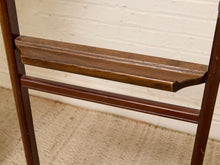 Load image into Gallery viewer, Close-up of a dark brown wooden chair or stool showing the backrest, a shelf, and a lower support rail against a white painted brick wall and beige woven rug.
