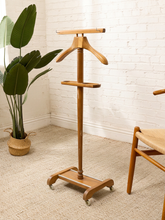 Load image into Gallery viewer, A mid-century modern wooden garment stand with a hanger and shelf, shown in a styled room corner with a white brick wall, beige carpet, a potted plant, and part of a wooden chair.
