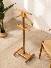 Load image into Gallery viewer, A light natural wood garment stand with a hanger, shoulder pad, and wheeled base, photographed in a styled room corner with a white brick wall, beige rug, potted plant, and part of a chair visible.
