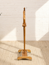 Load image into Gallery viewer, A freestanding wooden garment stand with a curved neck, multiple hook sections, and a wheeled base, photographed indoors against a white brick wall on a beige rug.
