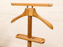 Load image into Gallery viewer, A vintage wooden garment hanger stand with a two-tiered design, featuring a curved wooden hanger and a lower flat tray, set against a white painted brick wall.
