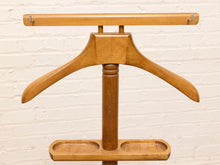Load image into Gallery viewer, A mid-century modern wooden hanger stand with a natural wood finish, featuring a top hanging bar, a central hanger, a vertical support column, and a bottom tray with two compartments, set against a white brick wall background.

